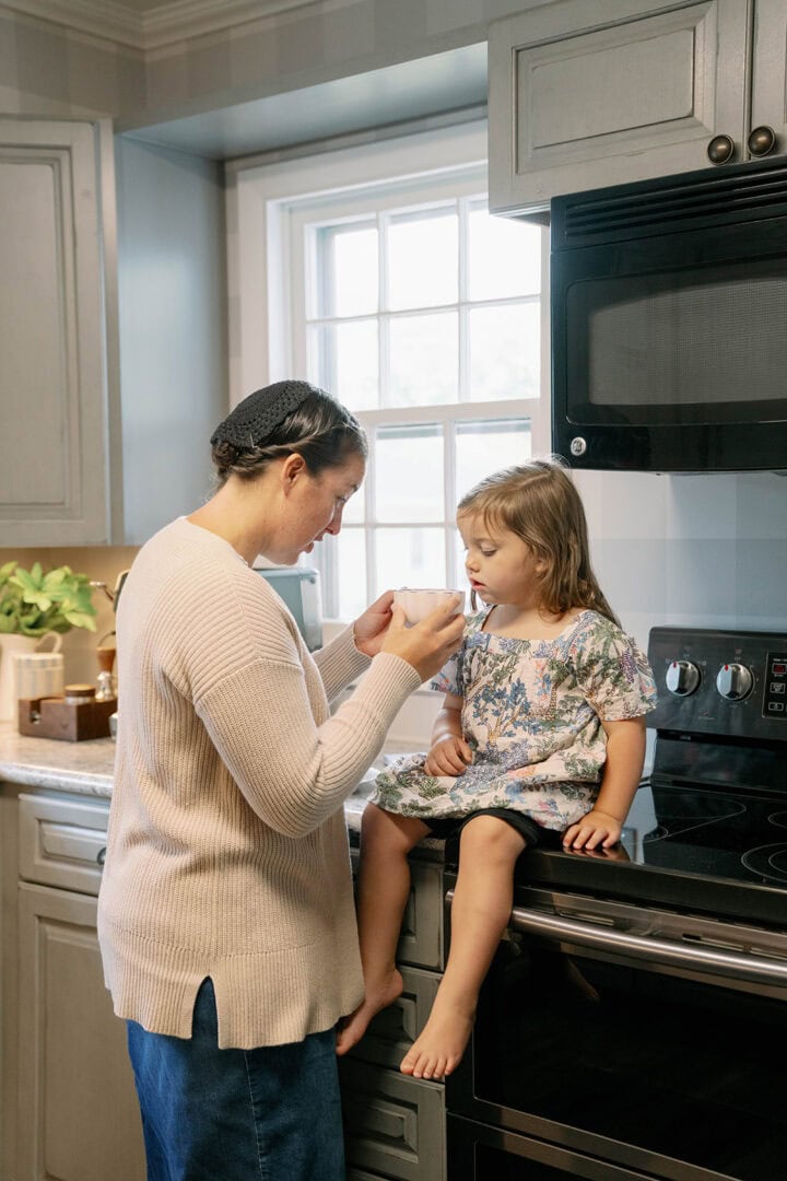 Mom giving hot chocolate to her three-year-old daughter in a kitchen with the toddler sitting on the countertop